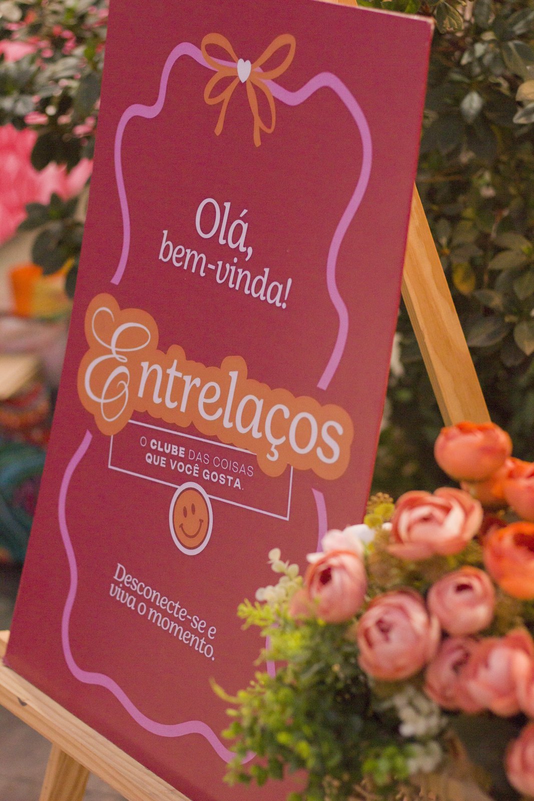 Placa do Entrelaços com a mensagem olá, bem-vinda, ao lado de flores
